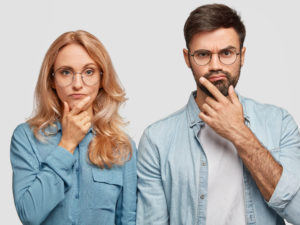 Horizontal shot of thoughtful male and female colleagues hold chins and being concentrated on solving problem, look directly into camera. Blonde middle aged female and her brother work as team